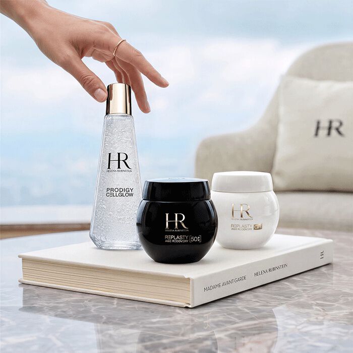 Tres productos de cuidado de la piel de Helena Rubinstein (Prodigy Cellglow y Re-Plasty) dispuestos sobre un libro blanco frente a una ventana con vistas al paisaje.​