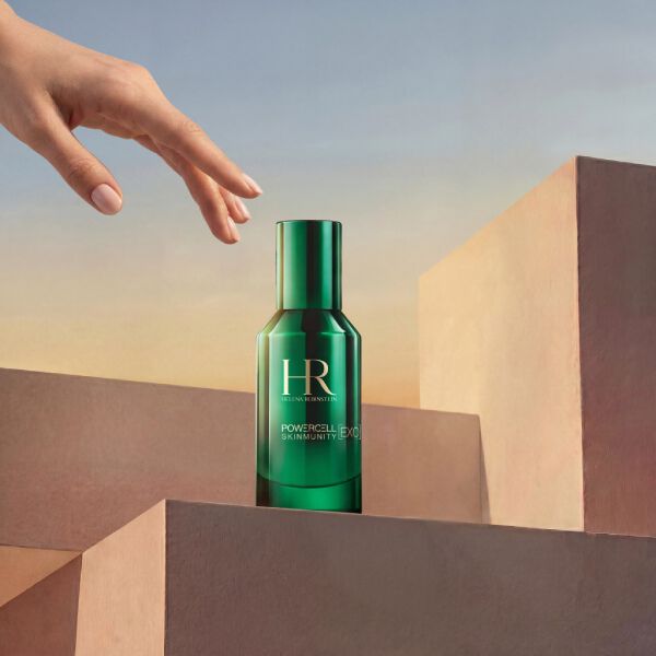 Foto del producto Serum Powercell Skinmunity Exo de Helena Rubinstein en un frasco de vidrio verde brillante. El frasco se encuentra sobre una pila de podios geometricos rosas, con una mano elegante que se acerca desde arriba, sobre un fondo de cielo con tonos calidos de atardecer.​