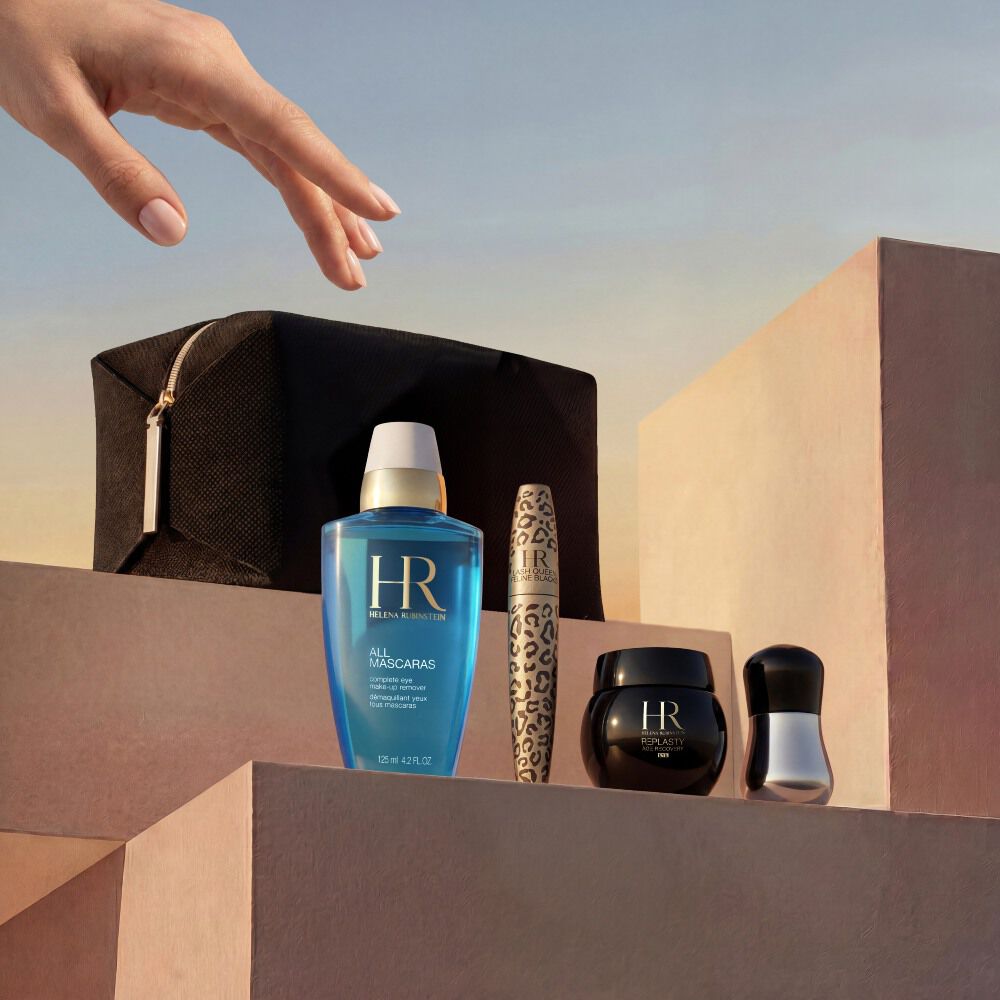  Una mano elegante se acerca a una seleccion de productos Helena Rubinstein dispuestos sobre bloques de piedra minimalistas en tono beige arena. El conjunto se presenta sobre un fondo de cielo dorado al atardecer e incluye un neceser de viaje negro, el desmaquillante de ojos azul 'All Mascaras', una mascara de pestanas Lash Queen Feline Blacks con estampado de leopardo, un pequeno tarro de crema Re-Plasty negro y una herramienta aplicadora plateada.​