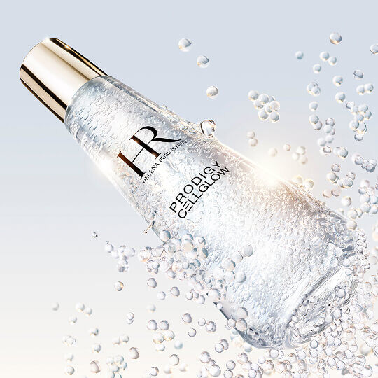 Frasco de suero Helena Rubinstein Prodigy Cellglow lleno de burbujas, inclinado y rodeado de burbujas brillantes suspendidas, sobre un fondo claro y luminoso.