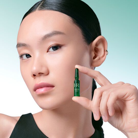 Primer plano del rostro de una mujer sosteniendo delicadamente entre sus dedos una ampolla verde Powercell [EXO] Skinmunity de Helena Rubinstein, sobre un fondo degradado en verde y blanco.​