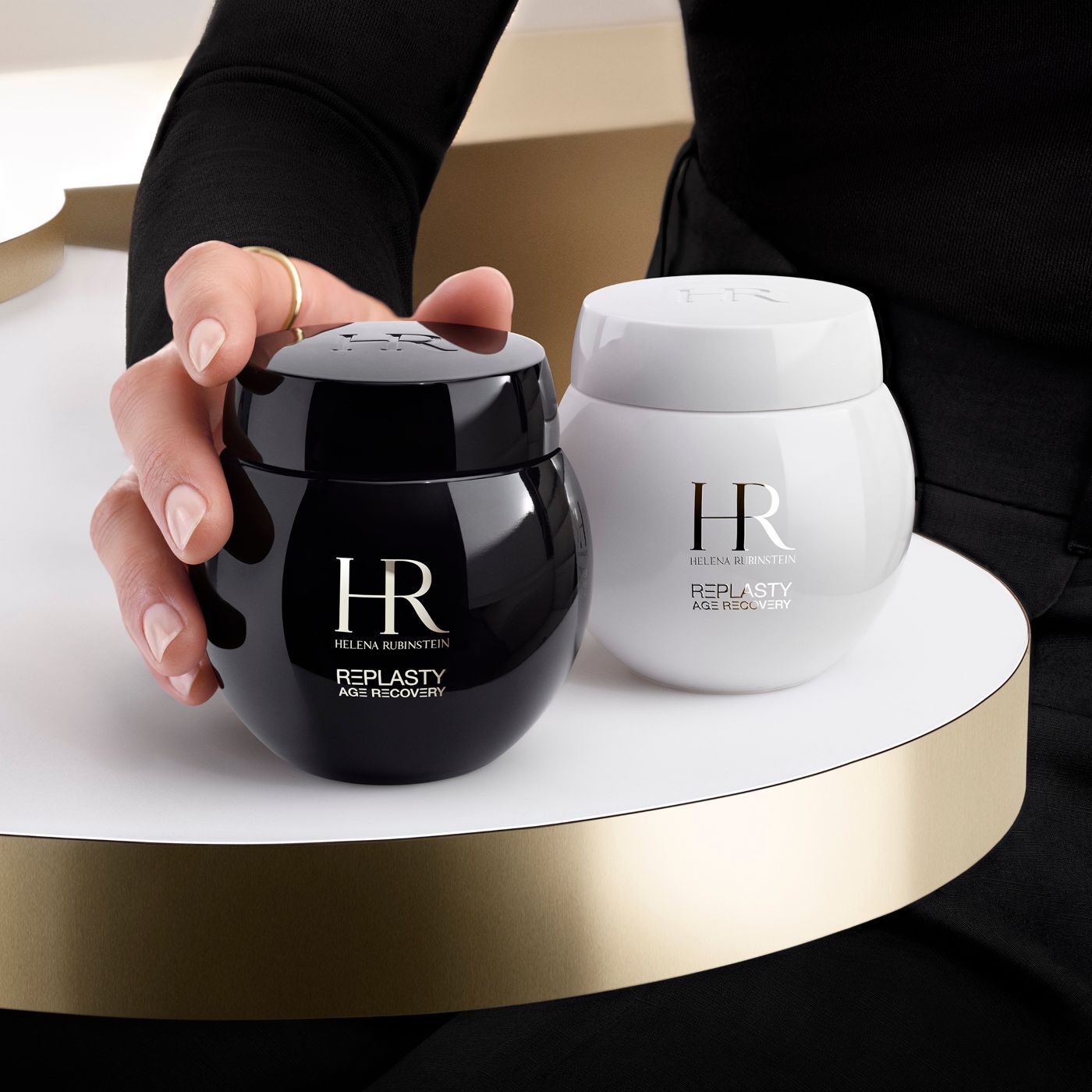 Una mano sujeta un tarro negro de crema Helena Rubinstein Re-Plasty Age Recovery, con un tarro blanco a juego al lado, sobre una plataforma dorada.