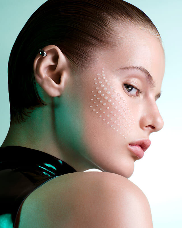 Mujer posando de perfil con gotas de serum en la mejilla sobre un fondo azul.