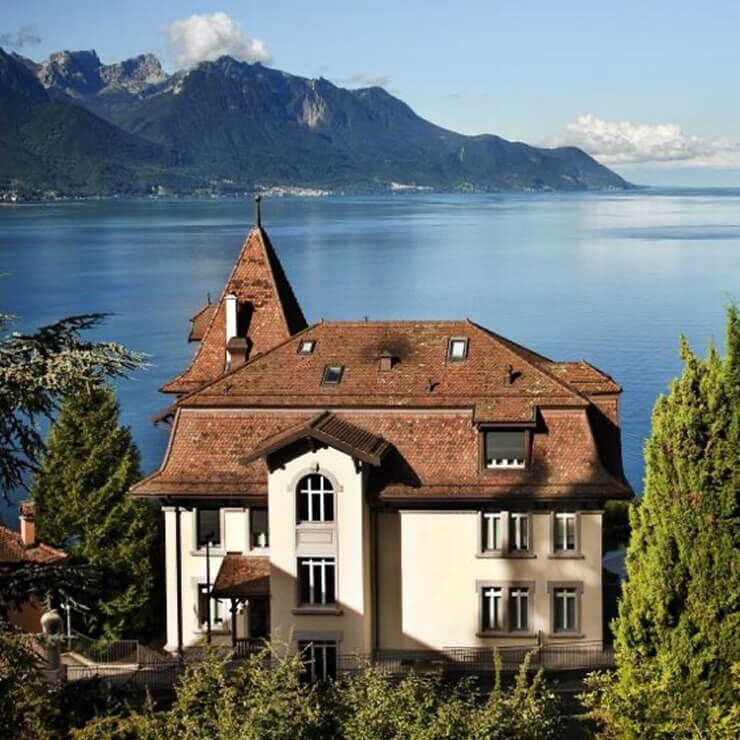 Una gran casa de estilo tradicional con tejado de tejas rojas y paredes claras, situada a orillas de un gran lago azul profundo, rodeada de montanas. El cielo esta despejado.