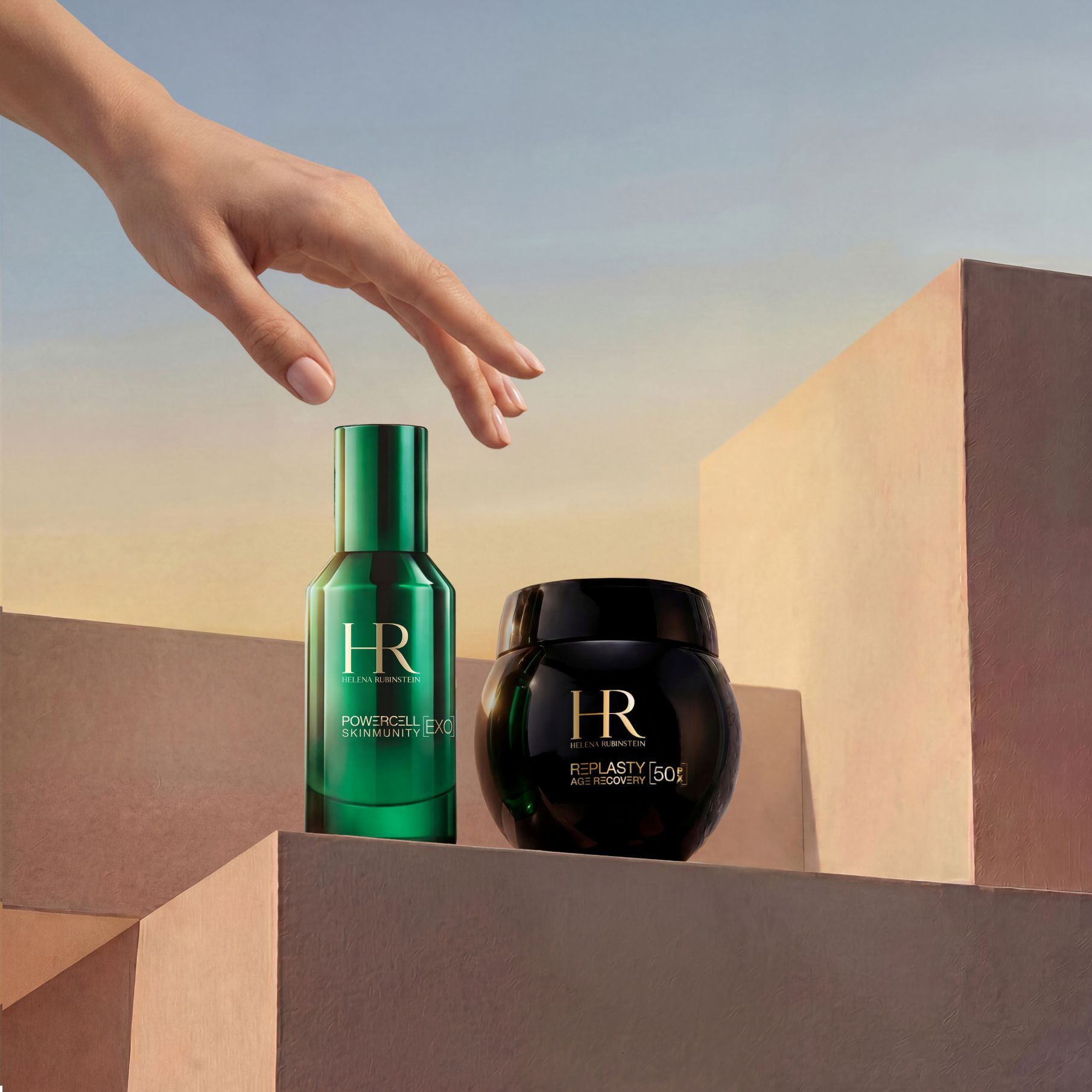 Una mano elegante alcanzando el Duo de Viaje Replasty Night – Powercell Essence de Helena Rubinstein, formado por un frasco verde de Powercell Skinmunity Essence y un tarro de Re Plasty Age Recovery Night Cream, presentados sobre bloques geometricos de arena con un calido cielo de atardecer de fondo.​
