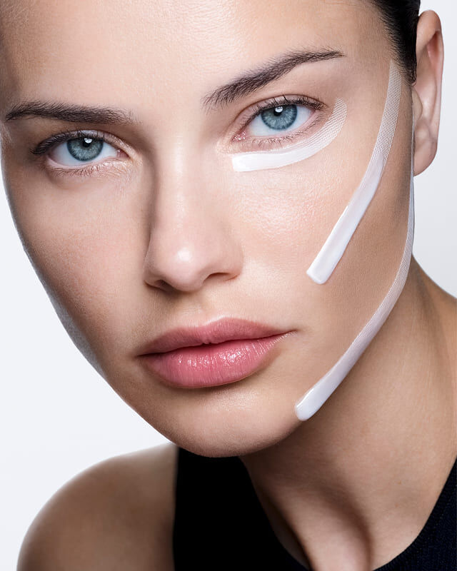 Adriana Lima con crema blanca y tiras aplicadas en el rostro, demostrando un efecto lifting visible.