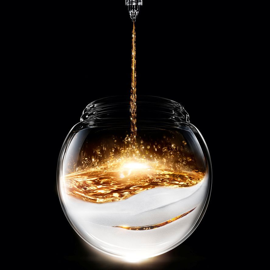 Liquido dorado brillante se vierte en un tarro de vidrio esferico, apoyado sobre una base blanca texturizada, creando un contraste luminoso sobre fondo negro.