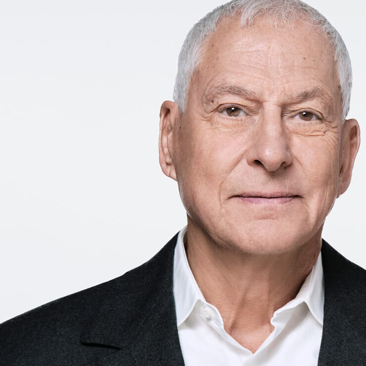 Primer plano de un hombre mayor con el pelo gris, camisa blanca y chaqueta oscura, mirando ligeramente de lado. Su expresion es neutra y su rostro esta iluminado.