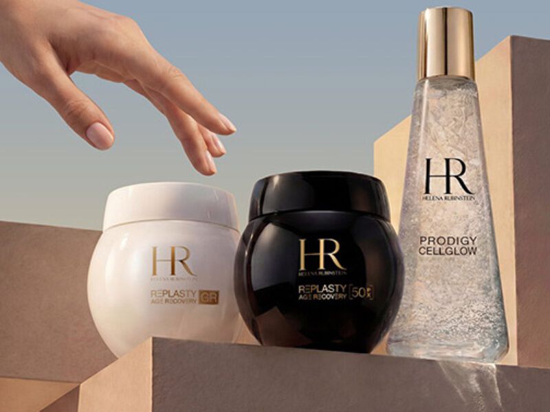 Una mano se extiende hacia tres productos de Helena Rubinstein, que destacan por su elegante diseno y su suave iluminacion.