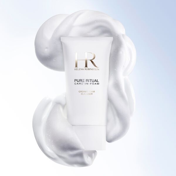 Primer plano del limpiador Pure Ritual Care-in-Foam de Helena Rubinstein rodeado de una espuma blanca y cremosa, junto al rostro de una mujer de tez pura y luminosa.​
