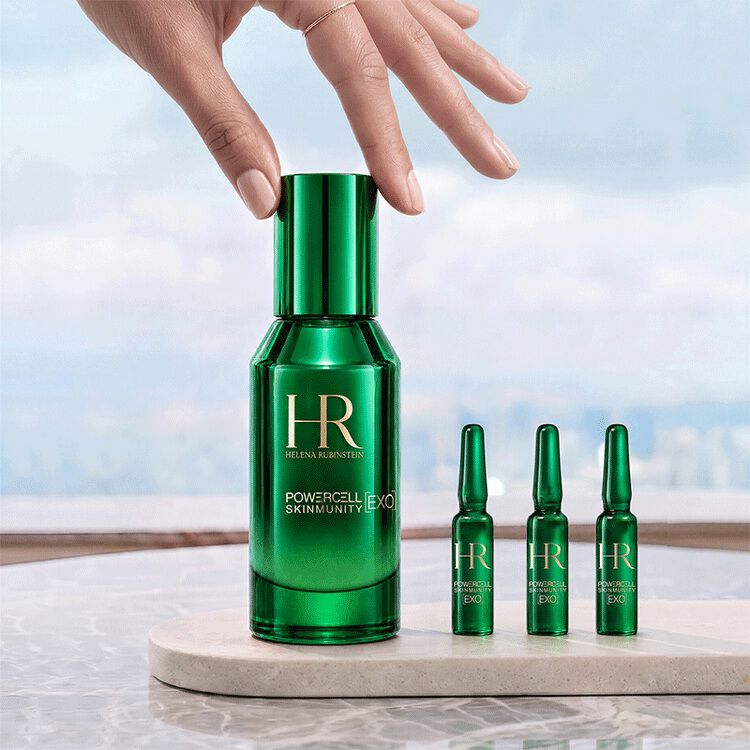 Una mano tocando el frasco verde del serum Powercell [EXO] Skinmunity de Helena Rubinstein, colocado junto a tres ampollas [EXO] Shots sobre un soporte de marmol frente a un paisaje luminoso.​
