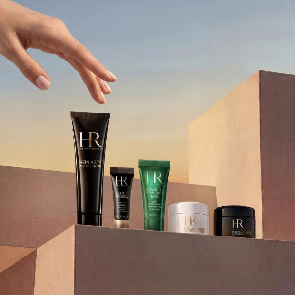 Fotografia de producto de la seleccion de regalos del mes de Helena Rubinstein. Bajo un suave cielo degradado, una mano delicada se desliza hacia tratamientos de lujo dispuestos sobre pedestales minimalistas color arena. Este exclusivo set incluye un frasco de Powercell Skinmunity, un tubo de viaje de Powercell Skin Rehab, dos tubos de viaje de Powercell Skinmunity Exo, un tarro de Re-Plasty Age Recovery Day Cream [GR] y un elegante neceser de viaje blanco de fondo.​
