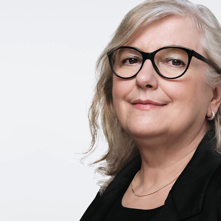 Primer plano de una mujer de mediana edad con cabello gris claro, gafas negras y un discreto collar dorado, sonriendo ligeramente.