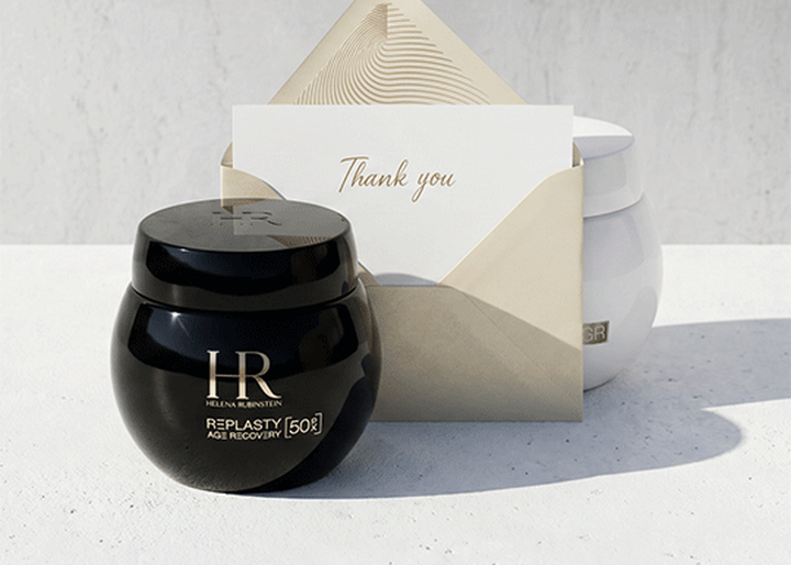 Un tarro negro de crema Helena Rubinstein Replasty Age Recovery y un sobre con una tarjeta de agradecimiento sobre una superficie blanca, con un tarro blanco al fondo.