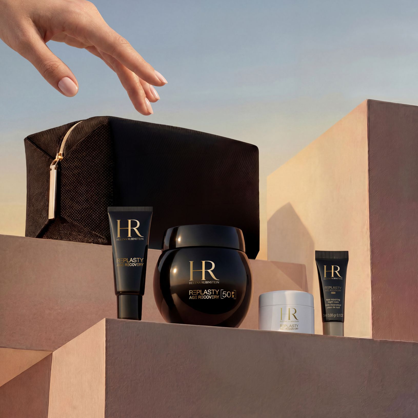 Una mano elegante se acerca a un set de regalo Helena Rubinstein, dispuesto sobre bloques de piedra color beige arena con un cielo de atardecer de fondo, que incluye un neceser de viaje de edicion limitada en negro y la gama Replasty Age Recovery: Eye Repairing Night Care, Day Cream, Night 50PX y Body Repairing Care.​