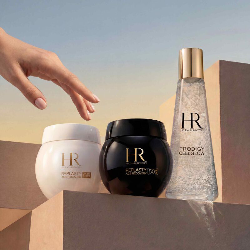 Frasco de serum Prodigy Cellglow (claro, burbujas) y tarro de crema Re-Plasty (negro) de Helena Rubinstein. Colocados sobre un fondo blanco texturizado y luminoso, simbolizando elegancia y eficacia del cuidado.